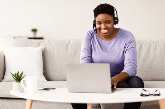 Telecommuting Concept. Smiling curvy african american woman sitting on the sofa and working online on laptop, wearing headset. Young black female using computer at home office, typing on keyboard