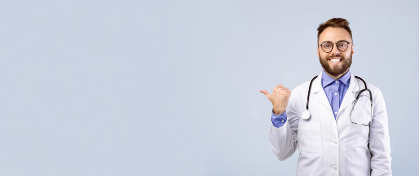 Positive Caucasian male doctor in lab coat showing thumb up gesture and pointing at empty space over grey studio background, banner design. Healthcare and medical concept