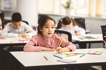 Tuition Concept. Portrait of concentrated small asian schoolgirl sitting at table in classroom, writing in copybook, listening to teacher and taking notes during lesson, looking at blackboard