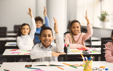School Reopening, Back To Normal Lifestyle Concept. Portrait of excited little schoolboy raising hand up in the air to answer a question with all his classmates in blurred background, selective focus