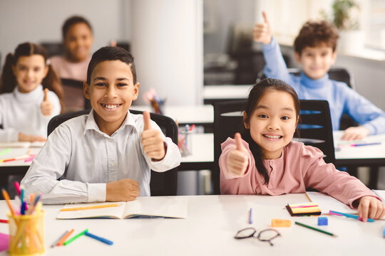 Education Concept. Diverse group of happy smiling international classmates sitting at desk in classroom and showing thumbs up sign gesture, girl and boy enjoying studying at school - Powered by Adobe