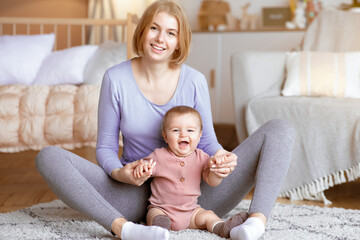 Obraz premium Happy beautiful caucasian mother holding laughing baby hands, playing with her cute infant child, sitting on floor in living room, copy space. Mom and baby having good time at home, playing together