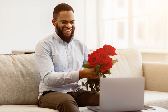 Virtual Dating During Pandemic. Smiling black man holding bouquet of red roses flowers and showing it to webcam, having online video call, sitting on couch. Long Distance Relationship - Powered by Adobe