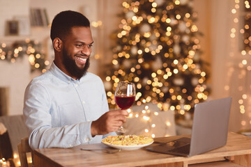 Virtual Meeting. Smiling african american man sitting at dining table, having dinner, holding glass of red wine, having video call with girlfriend, friends or family on laptop. Stay home