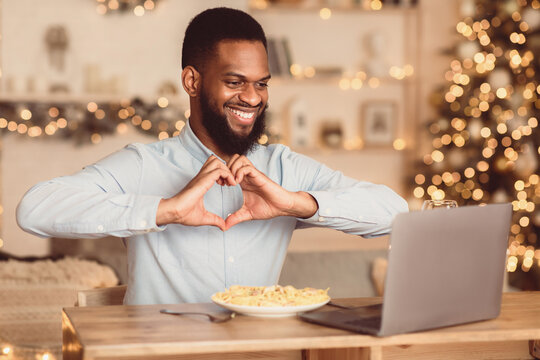 Love And Care. Portrait of smiling african american man making video conference while having dinner, showing heart sign gesture with hands to webcam. Guy sitting at dining table, eating pasta