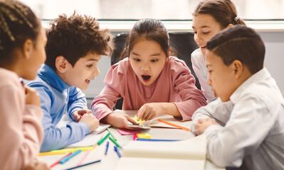 Children And Technology Concept. Diverse excited group of emotional happy junior school kids sitting at desk and using mobile phone, watching video during break. Kids and Gadget Addiction