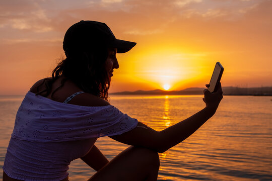 A girl taking a selfie on a smartphone, against the backdrop of a sea sunset on a beach. People vacation lifestyle journey concept.