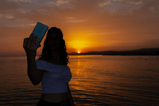 A girl taking a selfie on a smartphone,  against the backdrop of a sea sunset on a beach. People vacation lifestyle journey concept.