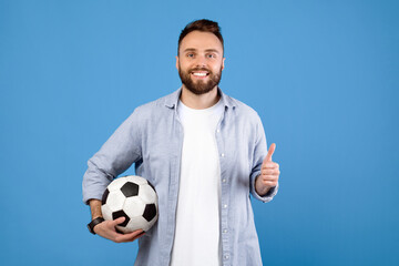 Cheerful young guy holding soccer ball, making thumb up gesture, recommending sports on blue studio background. Portrait of football fan supporting his favorite team during championship