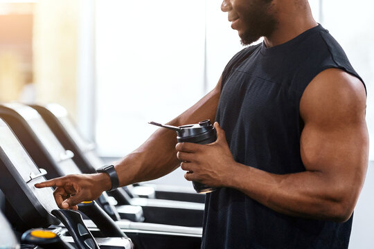 Sports drinks, nutrition concept. Side view of muscular african american man with protein drink standing on treadmill, copy space, sun flare. Athletic black guy exercising at gym, jogging