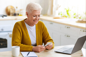 Modern technologies and old people concept. Happy senior woman reading messages while working on laptop at cozy kitchen at home, using mobile phone. Cheerful old lady using financial app on smartphone