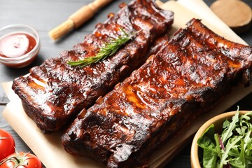 Tasty roasted pork ribs served with rosemary, tomatoes, sauce and salad on black wooden table, closeup