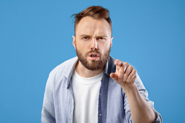 It is your fault. Angry Caucasian man pointing finger at screen in accusation on blue studio background. Frustrated young guy blaming you. Negative human emotions concept