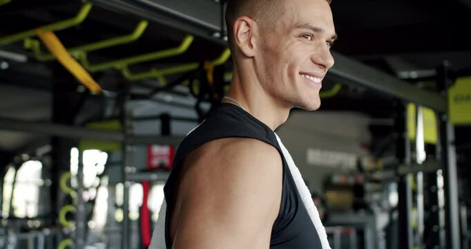 A fit man with a pleasant smile rests in a modern gym, towel draped over his shoulder, ready for his next workout. Smiling Fit Man Resting at Gym with Towel on Shoulder