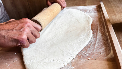 Close up of male's hands rolling dough on wooden table with wooden rolling pin. Homemade baking,...