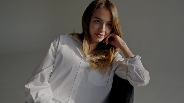 A portrait of a young woman wearing a white shirt, comfortably posing with her hand on her chin, illuminated by soft natural light. Young Woman in White Shirt Posing Casually in Soft Lighting