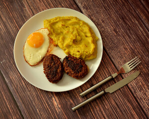 A large plate with a fried egg, mashed potatoes and two meat patties on a wooden table, cutlery nearby.