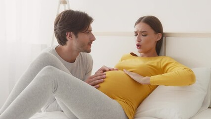 A man gently touches his partner's baby bump while pregnant woman feeling discomfort. The soft lighting creates an intimate atmosphere in their cozy bedroom - Powered by Adobe