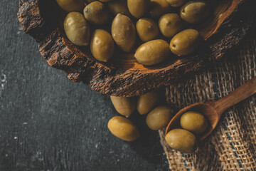 Rustic wooden bowl filled with green olives on burlap fabric with olive branch in background....