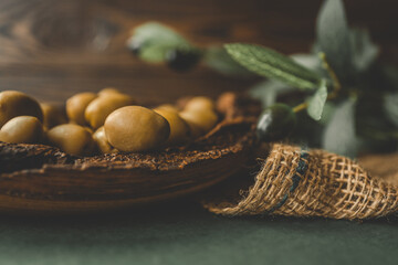 Rustic wooden bowl filled with green olives on burlap fabric with olive branch in background....