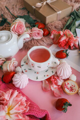 Flat lay of a cozy pink-themed setup with a gift box, flowers, candle, pink tea in a floral cup, white teapot, strawberries, and assorted sweets on a pink and fluffy background