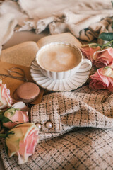 Warm cozy flat lay with a cup of frothy coffee, brown macaron, pink roses, a wrapped gift, and an open book, all placed on textured beige fabrics. Soft vintage morning vibe
