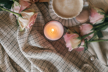 Warm cozy flat lay with a cup of frothy coffee, brown macaron, pink roses, a wrapped gift, and an open book, all placed on textured beige fabrics. Soft vintage morning vibe