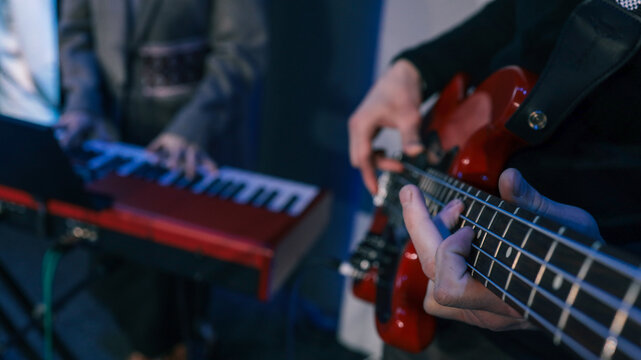 Close-up of hands playing electronic keyboard during live music performance with blurred bassist in background under blue lighting