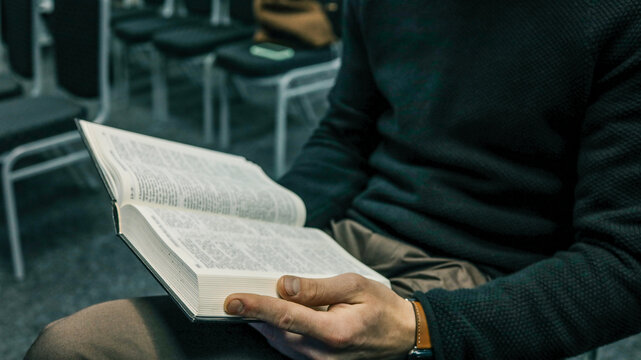 Close-up of a person holding an open Bible in their lap while sitting in a church auditorium with rows of empty chairs
