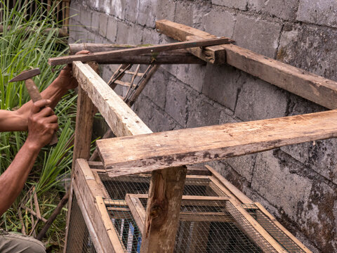 A man repairs a bamboo chicken cage with a hammer. The rustic setting highlights traditional craftsmanship and rural life.