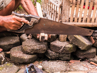 A man using a rusty handsaw to repair a bamboo cage on a stone floor. This scene captures...