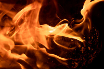 Close-up image of bright orange flames against a backdrop of black ash and burnt debris. A strong...