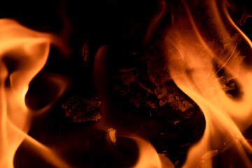 Close-up image of bright orange flames against a backdrop of black ash and burnt debris. A strong...