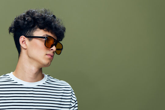 Young man with curly hair wearing sunglasses and a striped shirt posing against a solid olive green background in a casual style portrait. - Powered by Adobe