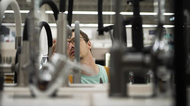 Young man wearing glasses carefully examining various faucets displayed in a hardware store, considering different models before making a purchase