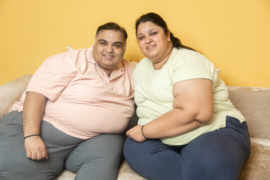 Fototapeta Smiling plus size indian couple looking at camera. Overweight husband wife sitting on couch. Fat chubby man and woman together. Obesity