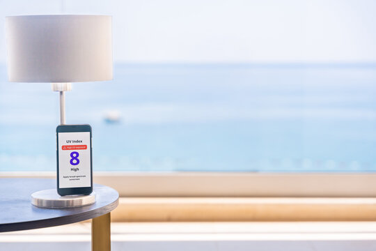 Smartphone displays high UV alert with sunny beach and sea in background. Sun safety reminder during seaside vacation