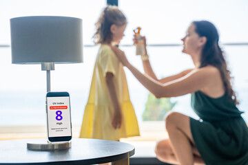 Smartphone with high UV alert in focus, while mother applies sunscreen to daughter in background....