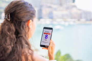Young woman checking UV index on smartphone with high radiation alert. Mobile app warns to apply...