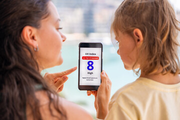 Mother holds smartphone with high UV alert while teaching daughter about sun safety. Educational...