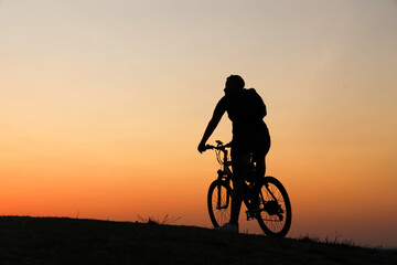 black silhouette of Young one alone male person man standing on hill horizon line with their bicycle and looking at far away on yellow orange sunset sky background. empty copy space for inscription