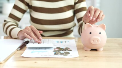 An Asian woman is putting money in a piggy bank while looking at a calculator, giving advice on financial planning. - Powered by Adobe
