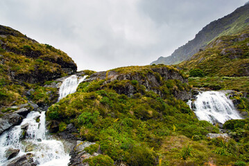 A breathtaking view of cascading waterfalls flowing over rugged, moss-covered cliffs along the iconic Routeburn Track in New Zealand. The lush green vegetation, misty atmosphere, and dramatic rocky te