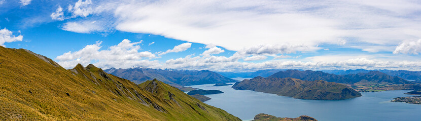 Breathtaking view  of a vast, deep blue lake nestled between rolling green hills and rugged mountain ranges. The sky above is a vibrant blue, dotted with soft, scattered clouds. Kepler Track, New Zeal