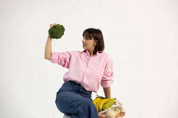 Smiling woman on white cyclorama holding broccoli and flexing biceps. Promoting healthy lifestyle,...