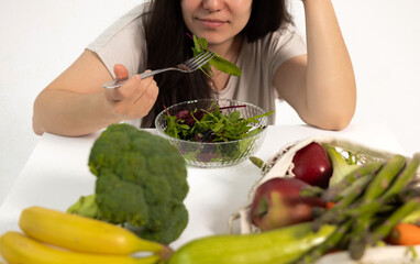 Woman surrounded by fresh vegetables eating salad with fork. Girl with extra fat holding body...