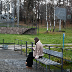 Fototapeta premium Young woman sitting alone on a bench in an empty park reading on a smartphone while surrounded by nature in early springtime