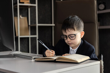 Funny baby boss in glasses sitting at desk with laptop, smartphone, and notebook. Child in suit...