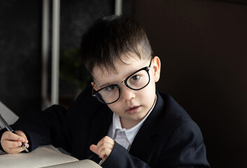 Funny baby boss in glasses sitting at desk with laptop, smartphone, and notebook. Child in suit...