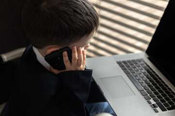 Funny baby boss in glasses sitting at desk with laptop, smartphone, and notebook. Child in suit...